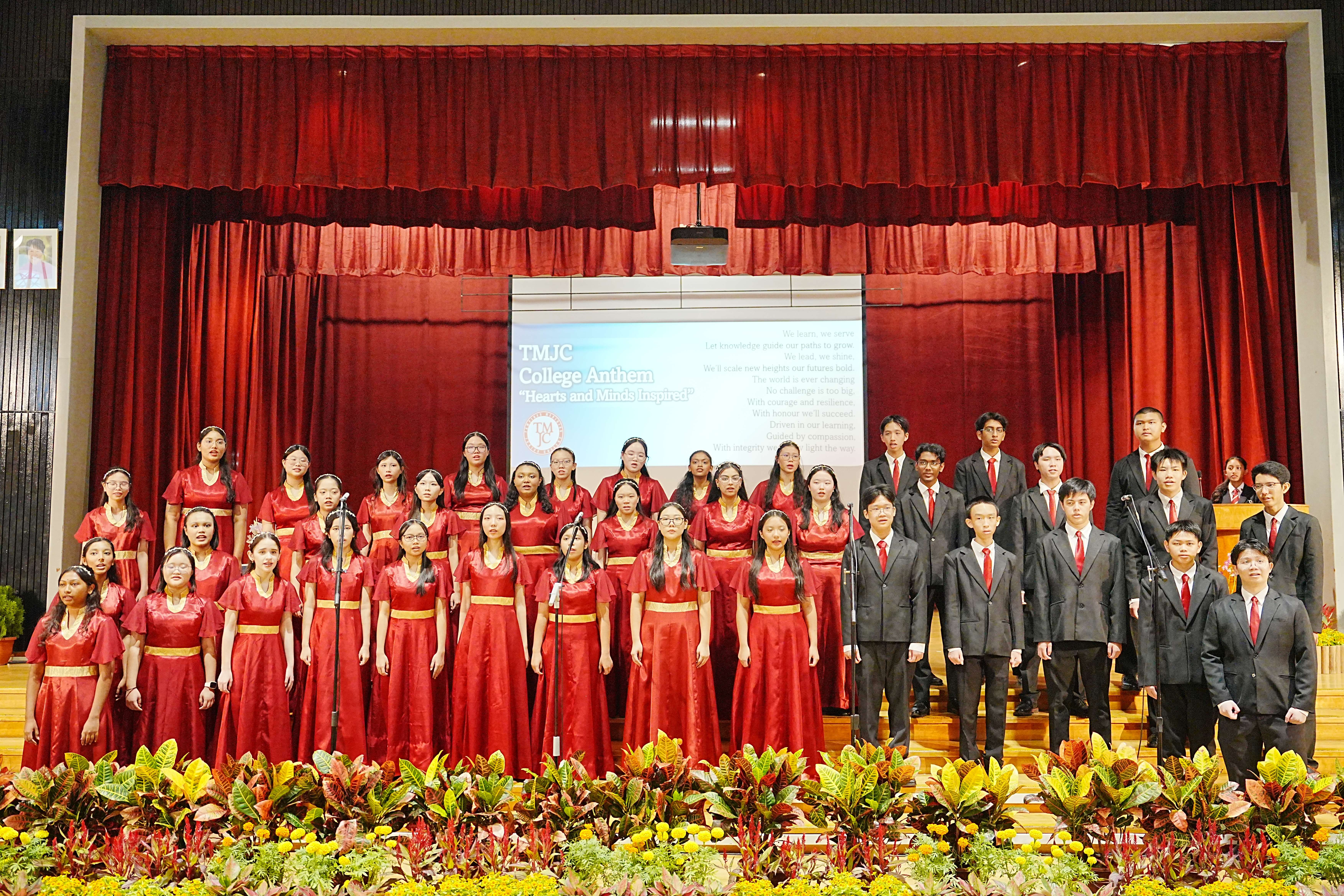 TMJC Choir performing the College Anthem 'Hearts and Minds Inspired', with female members in red gowns and male members in black suits.